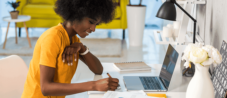 A black woman wearing an orange shirt is working on the computer in her home office.