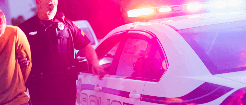 Policeman bringing a man in handcuffs to a police car with flashing lights.